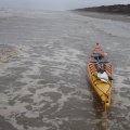 Вода оказывается не такой холодной и я в неопреновых тапочках вполне 
могу брести по щиколотку в воде с голыми ногами. Берег пологий и 
песчаный, лишь на мысах и в некоторых местах возникают выбросы камней и 
мне приходится уходить поглубже, чтобы пройти эти участки. Скорость 
буксировки  3-4 км/ч, при этом я устаю не очень сильно и не испытываю 
никаких переживаний. Во время буксировки все время замечаю высоту 
прилива и посматриваю вперед - берег обрывистый, и если начнет 
прижимать, лодку будет не затащить и придется выходить в море, а на 
приливе прибой заметно сильнее. Примерно через 1.5 часа, пройдя так 4.5 
км, я замечаю на берегу трапик, и чуть дальше лодку в море на привязи - 
значит тут есть люди. Решаю сделать остановку, тем более что дальше 
начинается большой мыс, который уже подперло водой и пройти его берегом я
 уже не смогу. Затащил лодку на берег, и пошел поискать людей. Здесь небольшой ручей и 
изба стоит на нем. В избе встретил Татьяну и Александра из Каменки. Они 
рассказали мне, что начался шторм, и его обещают на 4 дня. Начался он 
вчера, соответственно, сегодня и еще пару дней мне лучше стоять на 
берегу. Шторма здесь в это время редкость, а такой продолжительный - тем
 более. Но вот, мне не повезло. Оставаться мне здесь неудобно, поэтому я
 жду смены воды, чтобы продолжить путь. Сами они выехали собирать 
морошку, но ягоды в этом году нет, а тут еще и
 штормом прижало. Еды, конечно, взято надолго, а вот курева осталось на 
3-4 дня - проблема! Меня угощают обедом, пьем чай, общаемся - в основном
 Александр рассказывает мне о местных особенностях. Свою лодку они 
вдвоем вытащить на крутой берег не могут, поэтому сняли с нее все и 
оставили в море на якорях, там где меньше камней и ее не будет сильно 
бить. В самый прилив лодку все равно постепенно заливало водой, выбило 
стекло, но она еще держалась на плаву. Александр ждал отлива, чтобы 
пройти к лодке и откачать воду.  Дождавшись, когда вода немного убыла, я собрался с духом и вышел в море.
 Прибой прошел уже опробованным способом и отправился дальше - искать 
избу для штормовки. Александр мне рассказал, что дальше по берегу будут 
стоять на 2х катерах их знакомые, а потом будет еще много свободных изб.
 Здесь не побережье кроме рыбацких много изб морошечников (если есть 
смысл делать такое разделение), но сейчас многие из них уже пустуют. 